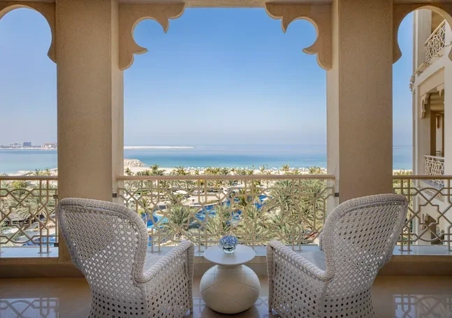 Waldorf Astoria Ras Al Khaimah, Al Hamra, Ras Al Khaimah: Resort balcony with ornate arched columns and a decorative railing, featuring two white woven armchairs and a small round table, looking out over palm trees, a swimming pool, a sandy beach,