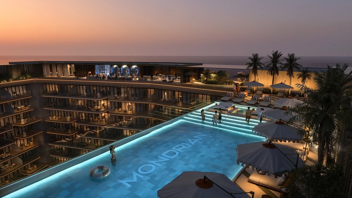 Resort swimming pool lit turquoise at twilight with the word Mondrian spelled on the pool floor, surrounded by grey cabana umbrellas, sun loungers, palm trees, and a curved multi-story hotel with balconies overlooking the sea.
