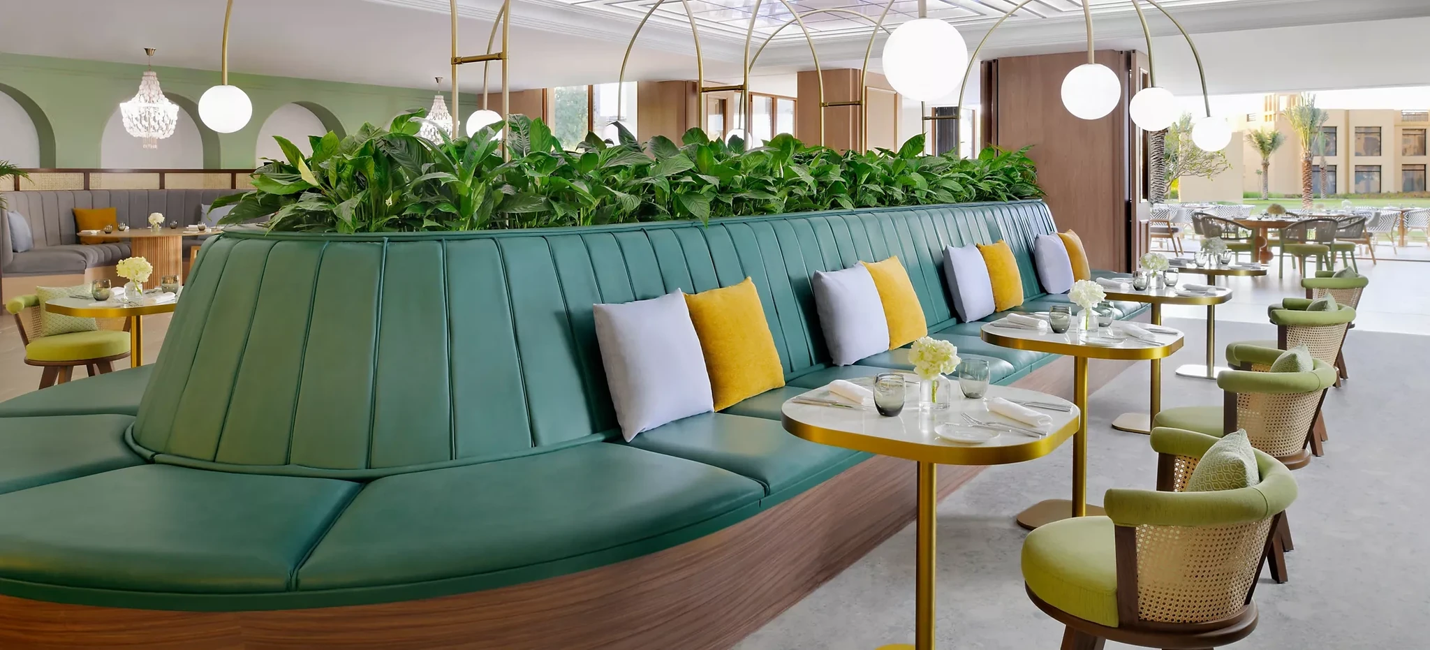 Restaurant interior featuring a long curved teal leather banquette with white and yellow cushions, a row of green plants above it, gold-edged marble tables set with glassware and flowers, green cane-back chairs, and globe pendant lights near arched windows.
