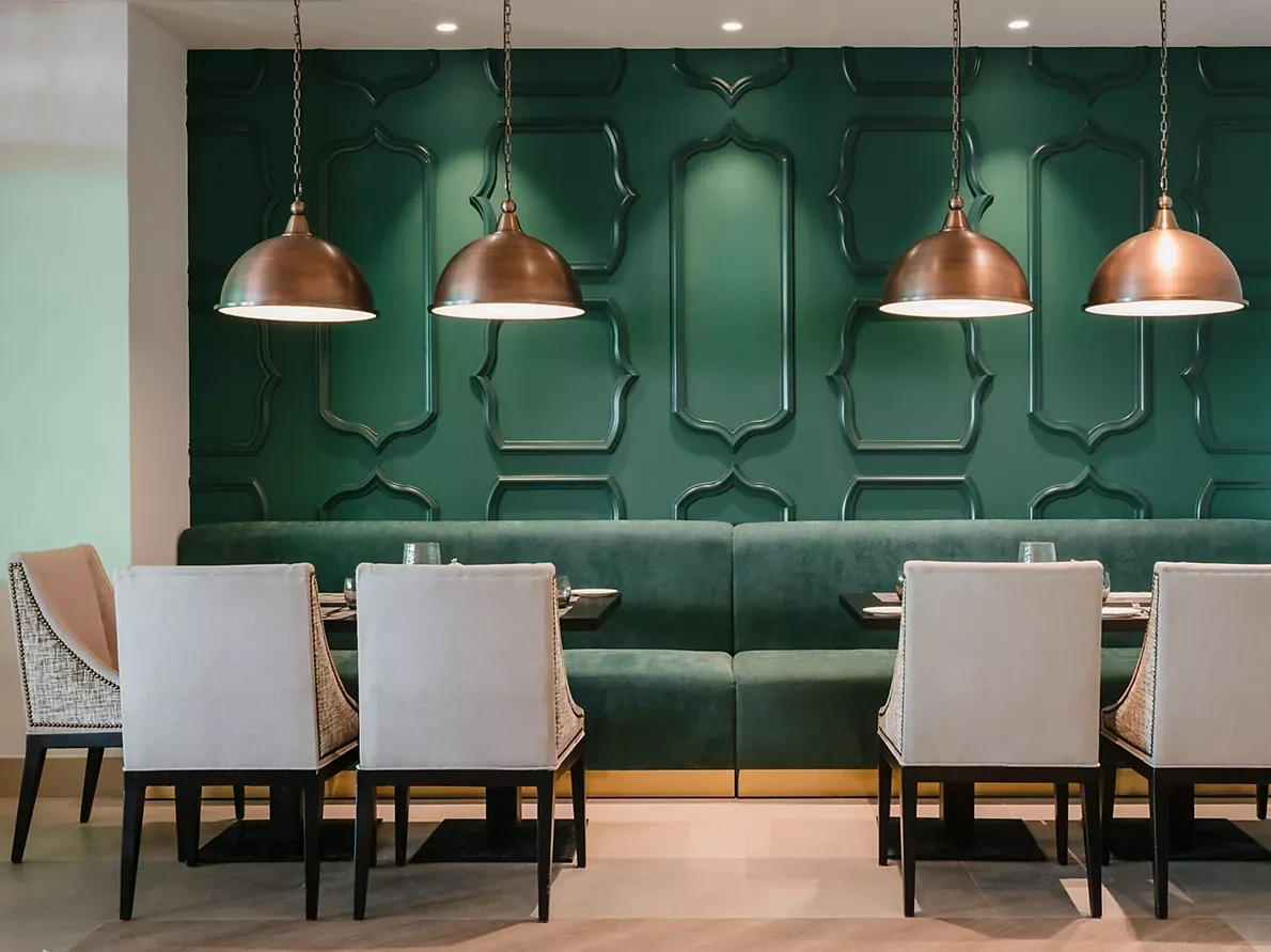 Restaurant interior with a dark green banquette bench along a patterned green panelled wall, cream upholstered dining chairs, set tables, and four copper dome pendant lights hanging from the ceiling.