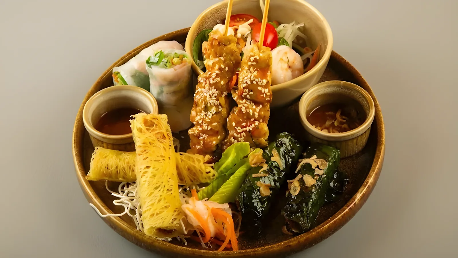 Round earthenware plate holding sesame-coated meat skewers, fresh rice paper spring rolls, crispy golden noodle nests, grilled leaf-wrapped parcels, and two small bowls of dark dipping sauce, over a gray background.