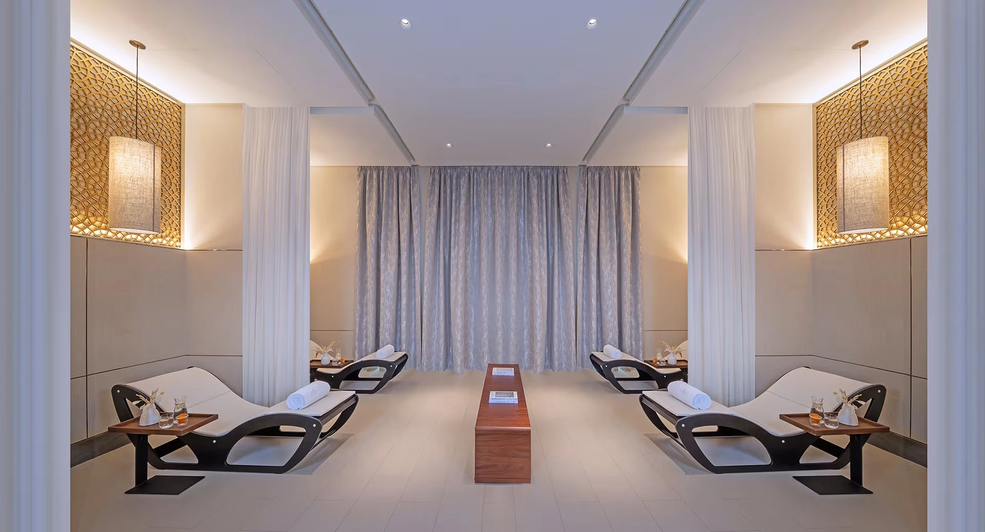 Anantara Mina Ras Al Khaimah Resort, Mina Al Arab, Ras Al Khaimah: Spa relaxation lounge with four black and white curved chaise loungers arranged in pairs, wooden side tables holding rolled towels and drinks, sheer curtains, a central wooden bench,