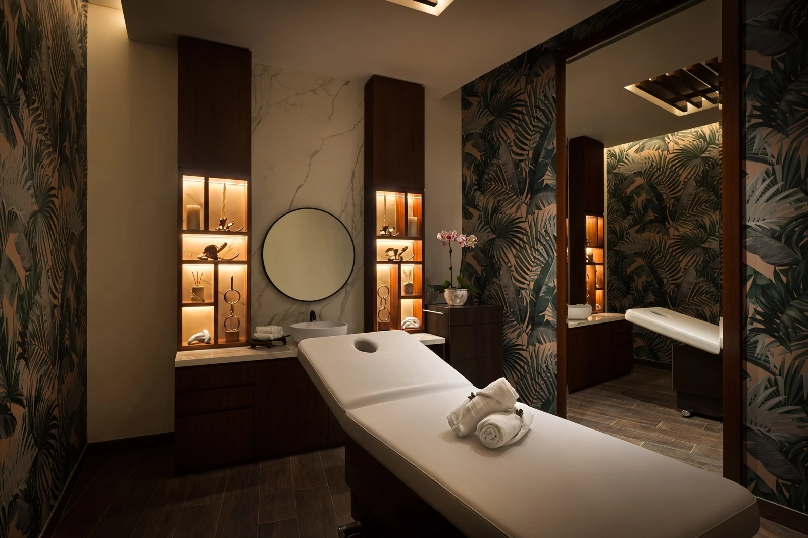Spa treatment room with a white massage table topped with two rolled towels, backlit wooden shelves holding candles and decor, a round mirror above a marble counter, an orchid, and dark green tropical palm leaf wallpaper.