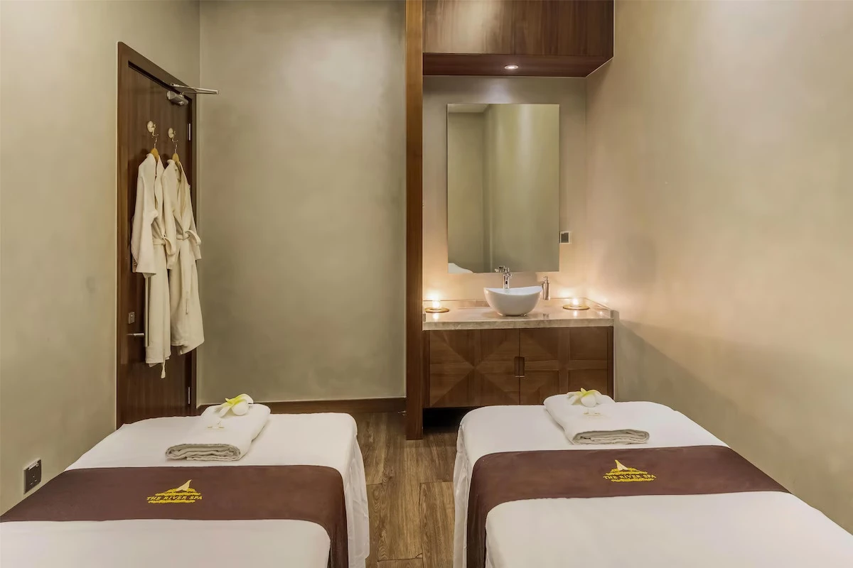Radisson Resort Ras Al Khaimah Marjan Island, Al Hamra, Ras Al Khaimah: Spa treatment room with two side-by-side massage tables dressed in white linens and brown runners, folded towels and white flowers on each bed, a wall mirror above a vessel sink