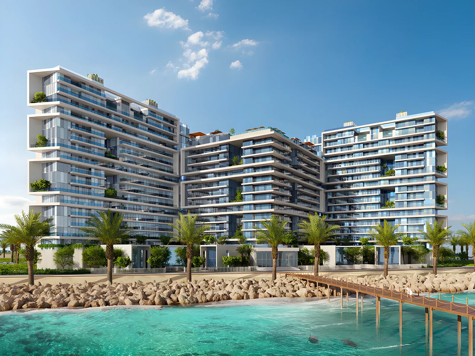 Three contemporary multi-story residential towers with glass balconies and terraces, fronted by palm trees, a rock breakwater, a wooden pier, and clear turquoise sea under a blue sky.
