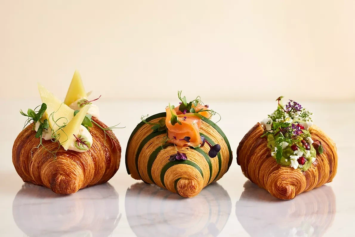 Three flaky croissants on a light marble surface, one topped with cream and yellow cheese shavings, one wrapped in green with smoked salmon, and one with avocado, crumbled cheese, pomegranate seeds, and microgreens.