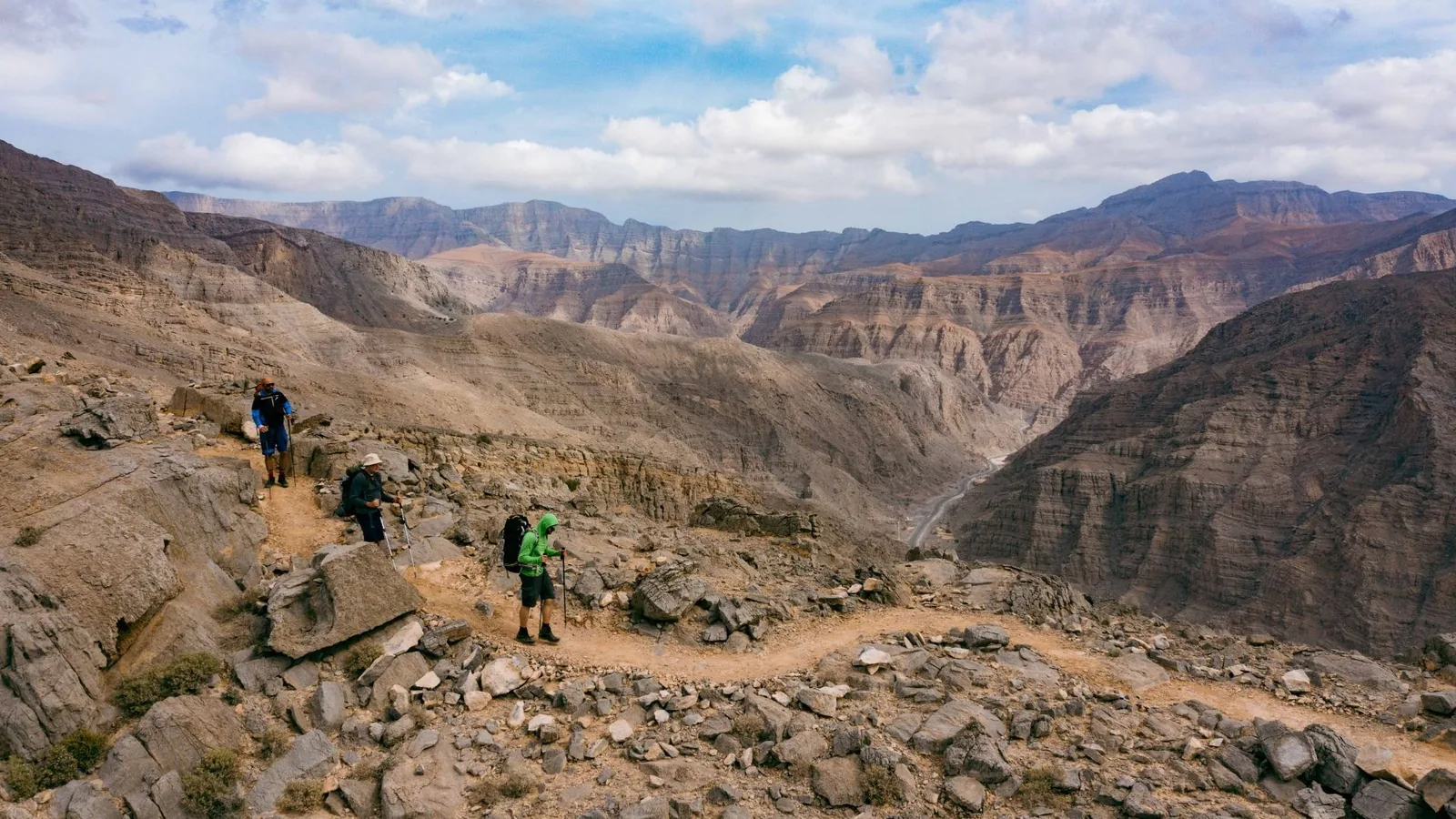 Three hikers on a stony trail high in rugged, layered mountains with a deep canyon and winding road below under a partly cloudy sky.
