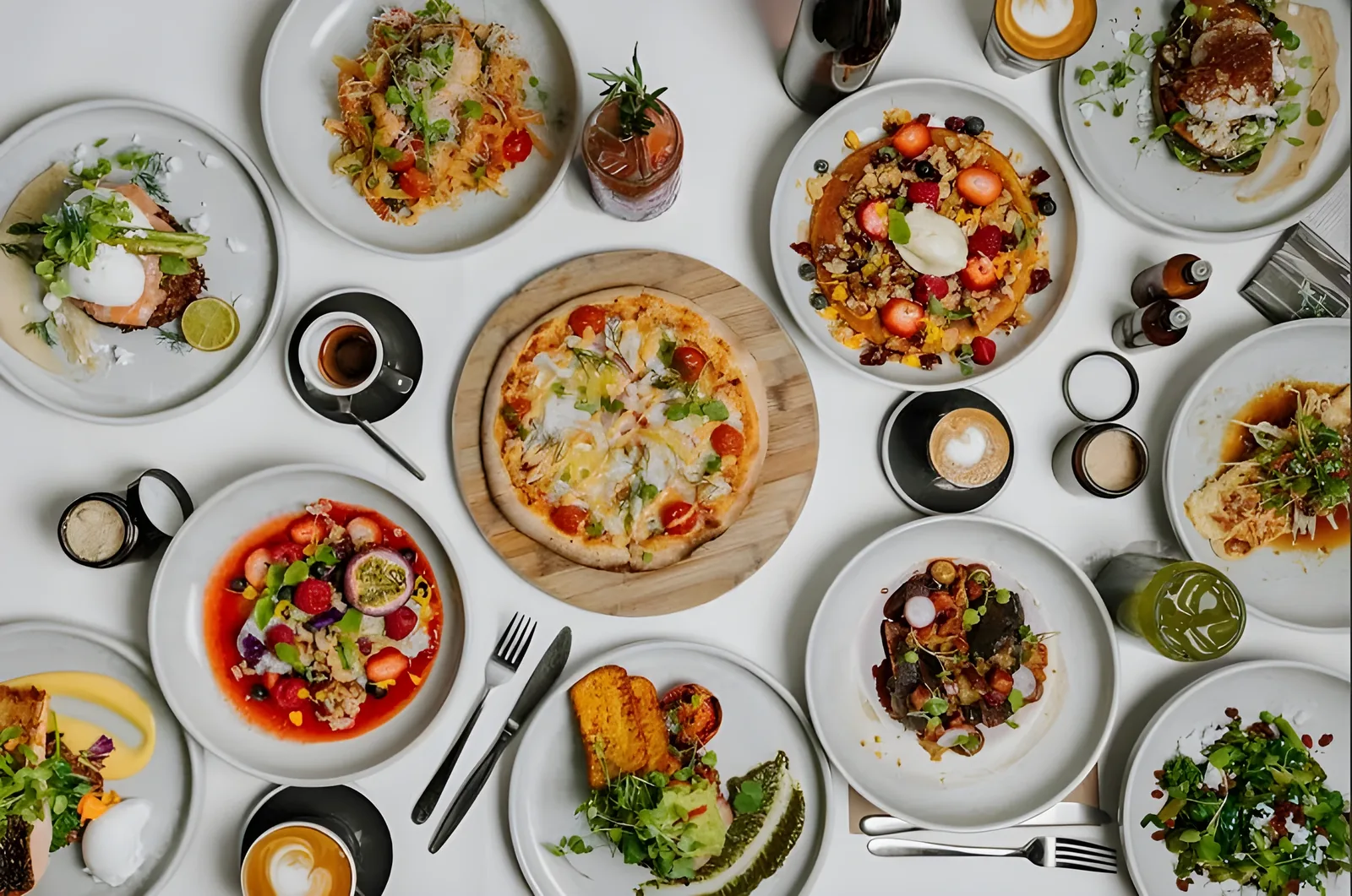 Top-down view of a white table set with many plates including a tomato pizza on a wooden board, salads, avocado toast, fruit dishes, cappuccinos in dark cups, and a green drink, with forks and knives scattered between them.