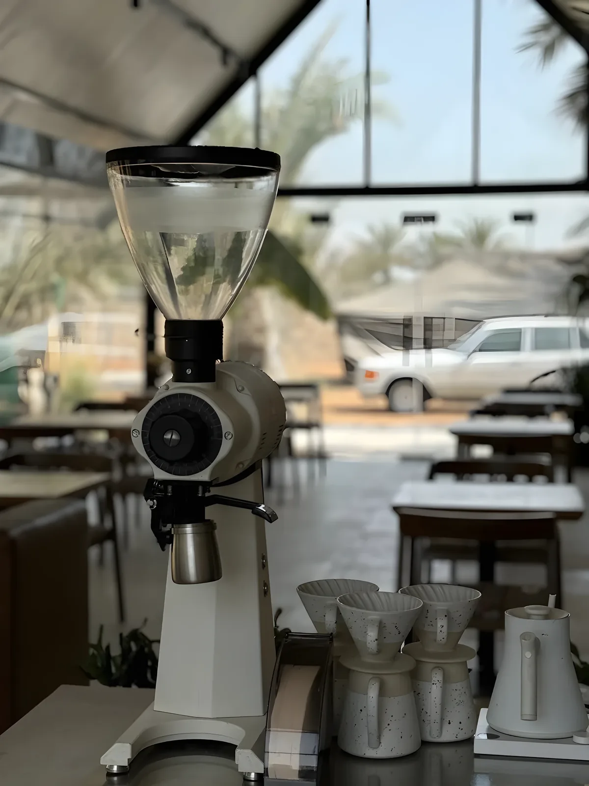 White espresso coffee grinder with a clear bean hopper on a cafe counter, next to speckled ceramic pour-over drippers and a kettle, with empty wooden tables and palm trees seen through glass windows in the background.