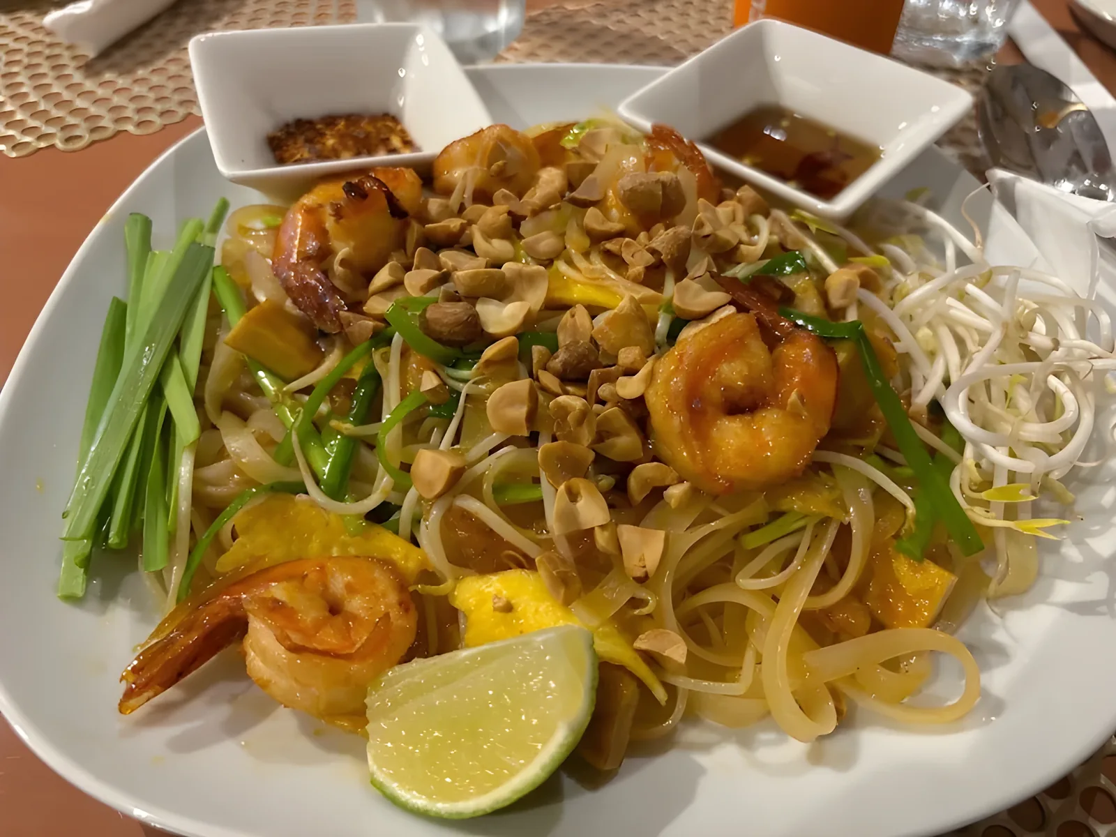 White plate of stir-fried rice noodles topped with grilled shrimp, crushed peanuts, bean sprouts, and green onion stalks, with a lime wedge and two square bowls of sauce on a dark table.