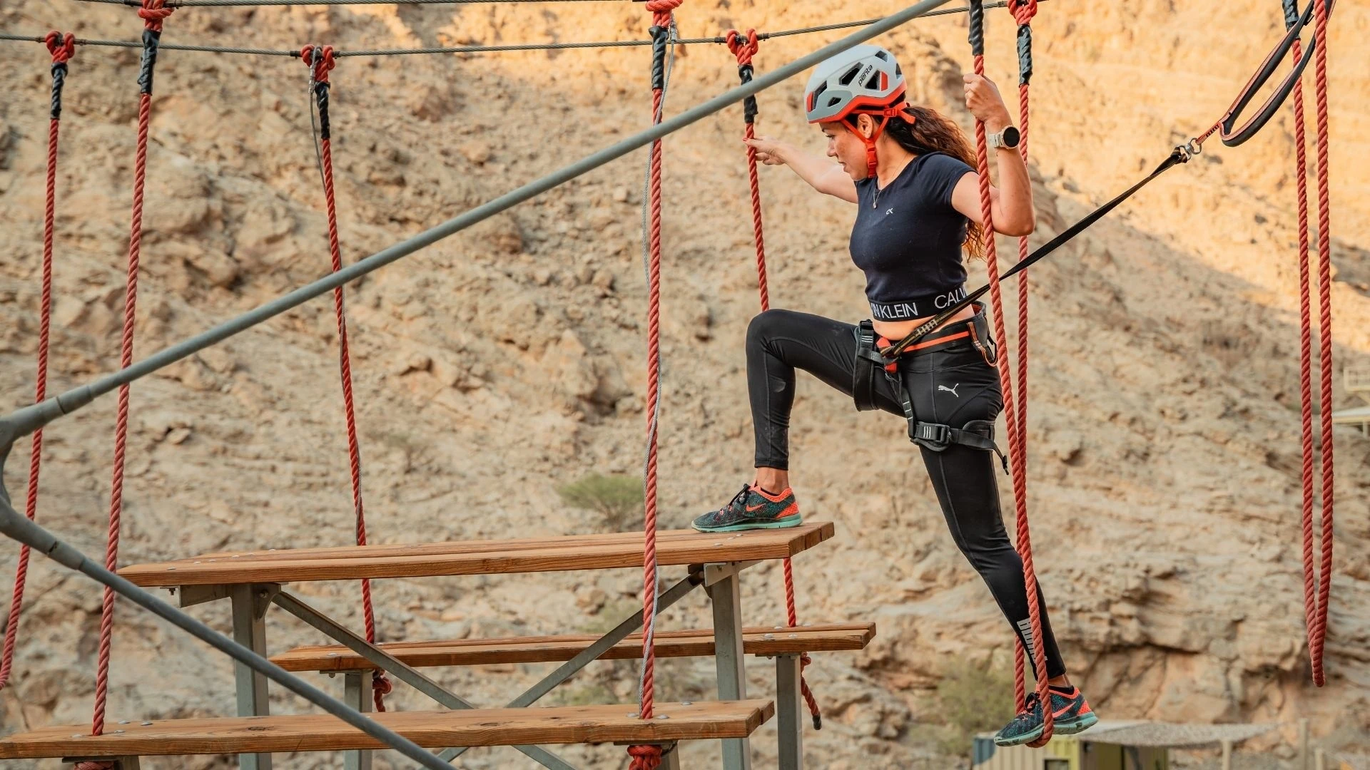 Jais Ropes Course Ras Al Khaimah, Ras Al Khaimah: Woman in a white helmet, dark top, and black leggings navigating a high ropes course, gripping red ropes with one foot on a wooden step and a rugged rocky mountainside behind her.
