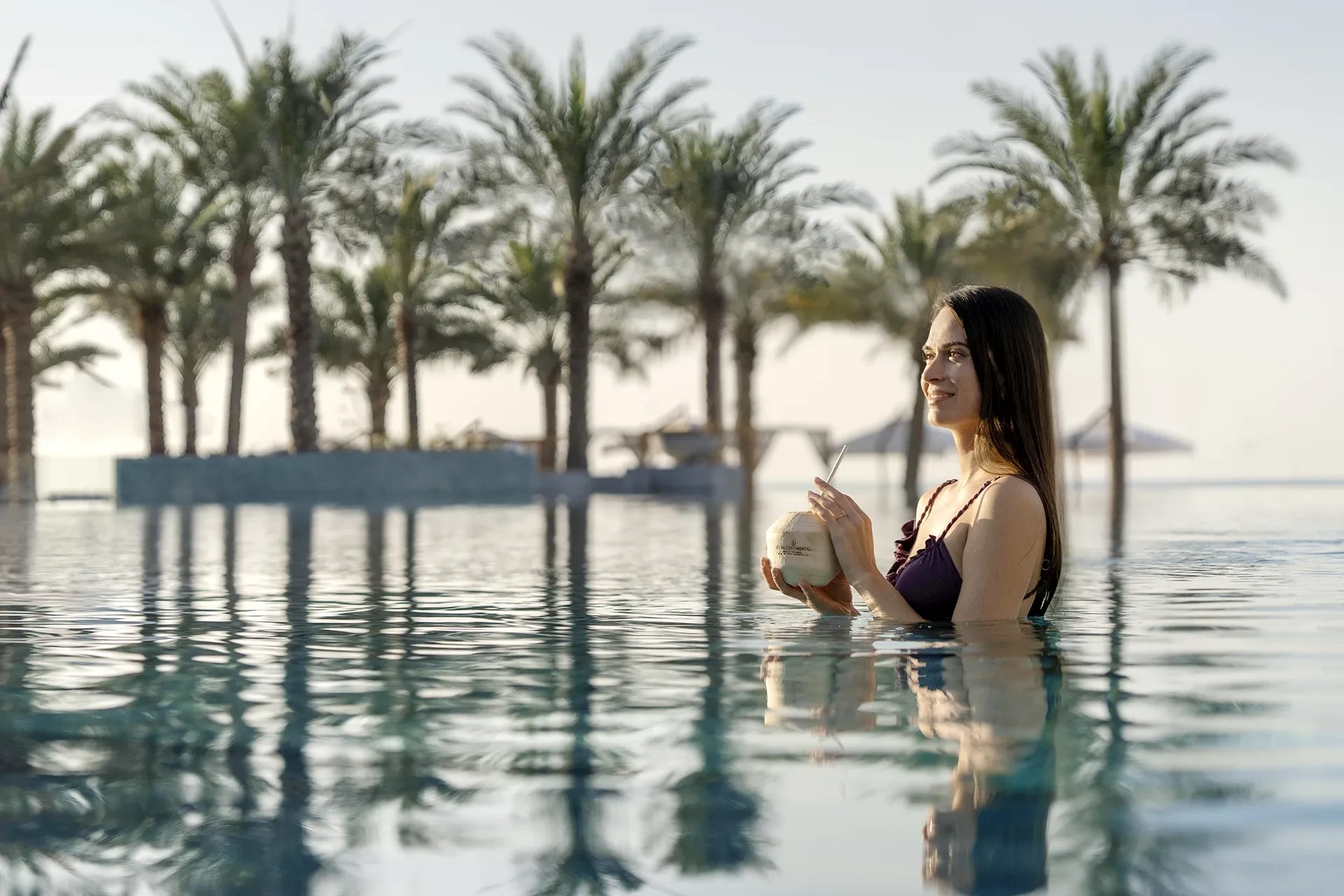 InterContinental Ras Al Khaimah Mina Al Arab Resort & Spa, an IHG Hotel, Mina Al Arab, Ras Al Khaimah: Woman with long dark hair in a purple swimsuit standing in an infinity pool, holding a coconut drink with a straw, palm trees and parasols behind