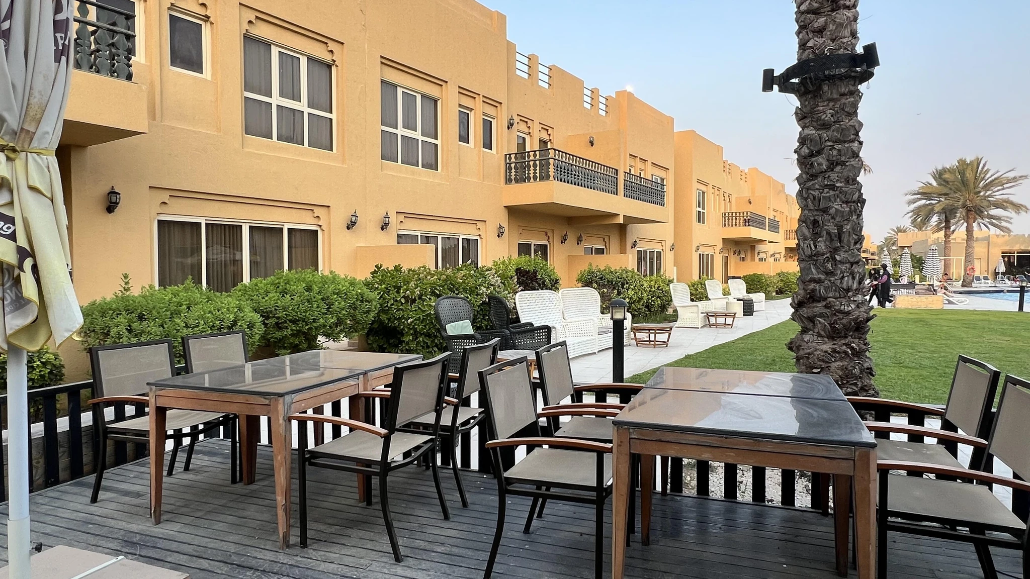 Wooden deck with glass-topped tables and grey mesh chairs on a resort terrace, backed by trimmed green hedges, a tall palm trunk, sand-colored two-story villas with balconies, and a pool area in the distance under a clear sky.