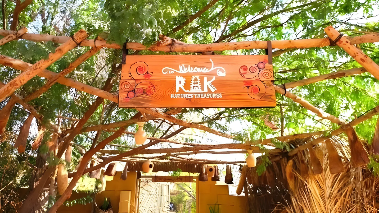Wooden hand-painted sign that says Welcome to RAK Nature's Treasures suspended from a rustic wooden pergola framed by green trees, dried gourds, and palm thatch.