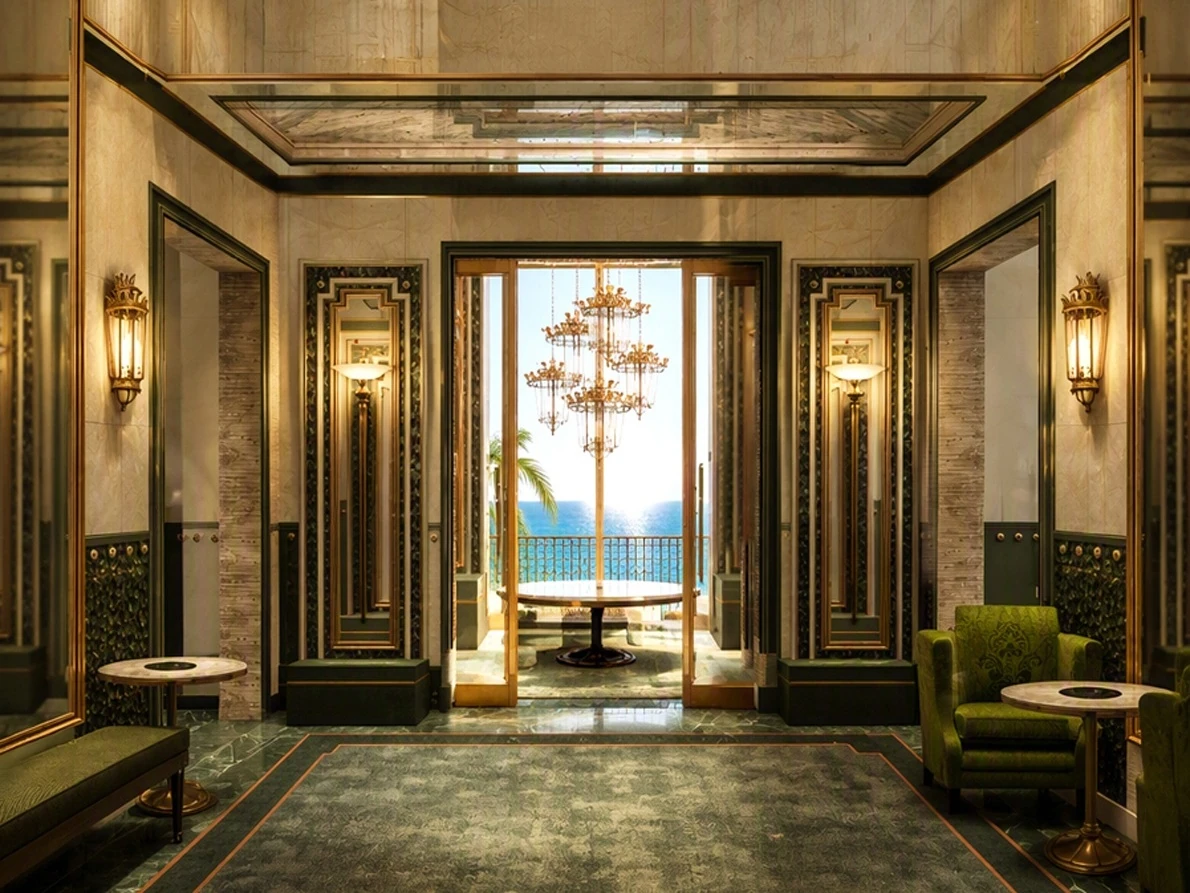 Opulent two-story hotel lobby with cream marble walls, brass wall sconces, ornate crystal chandeliers, a green marble floor, and green armchairs, with open double doors leading to a table and a sea view beyond.